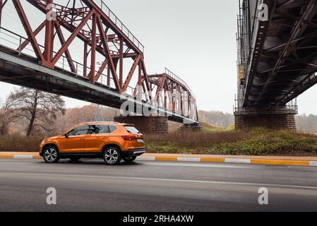 Vista posteriore di tre quarti del crossover samoa Orange SEAT Ateca. Riduzione della vista prospettica di due vecchi ponti a capriata sullo sfondo. Foto Stock