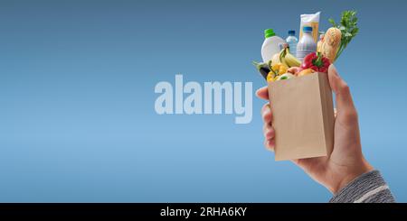 Cliente che tiene una borsa di carta in miniatura con alimentari freschi che cadono all'interno, concetto di shopping della drogheria Foto Stock