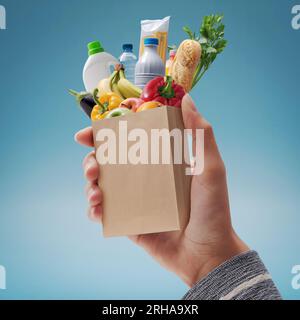 Cliente che tiene una borsa di carta in miniatura con alimentari freschi che cadono all'interno, concetto di shopping della drogheria Foto Stock