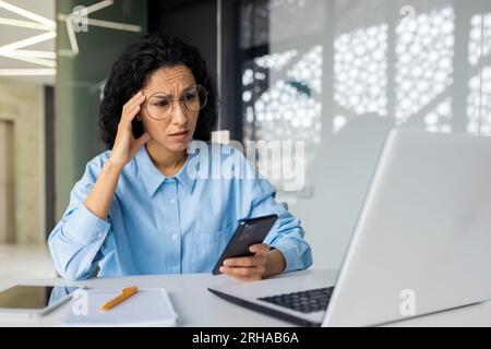 Triste donna d'affari sconvolta sul posto di lavoro, donna ispanica delusa dai risultati di lavoro all'interno dell'ufficio, lavoratrice che usa un computer portatile, che tiene in mano uno smartphone. Foto Stock