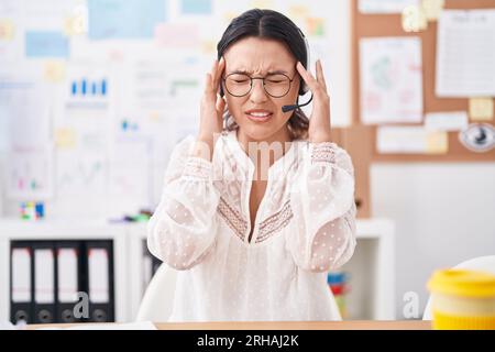 Giovane donna ispanica che lavora in ufficio, indossa cuffie e occhiali con la mano sulla testa, mal di testa per lo stress. soffre di emicrania. Foto Stock