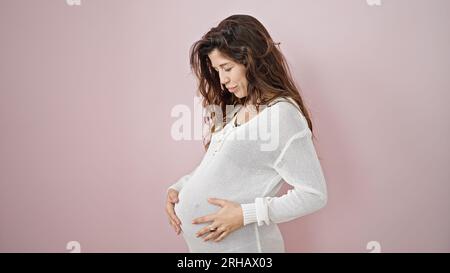 Giovane donna incinta in piedi con espressione seria che tocca la pancia su sfondo rosa isolato Foto Stock
