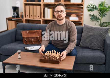 Uomo caucasico di mezza età che gioca a scacchi seduto sul divano pensando all'atteggiamento e all'espressione sobria che sembra sicuro di sé Foto Stock