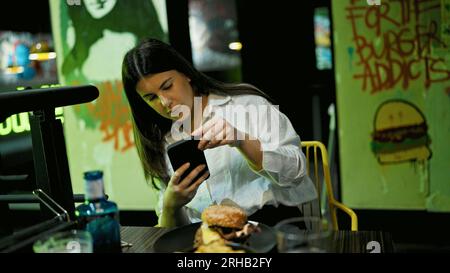Giovane bella donna ispanica che scatta foto di deliziosi hamburger con smartphone al ristorante Foto Stock