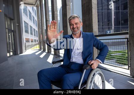 Uomo disabile dai capelli grigi che indossa una tuta ufficiale in sedia a rotelle, solleva il palmo, dà cinque in alto Foto Stock