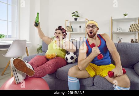 Donna grassa divertente con cibo e birra e uomo in forma con manichino che guarda il calcio in TV Foto Stock