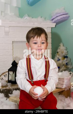 Un bambino vestito di rosso si diverte nella neve artificiale accanto al camino dello studio. Un ragazzino in costume rosso di natale che si diverte accanto a un Foto Stock