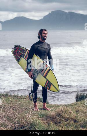Tutto il corpo di un giovane ragazzo ispanico barbuto con capelli scuri bagnati in muta che regge la tavola da surf e sorride dopo aver cavalcato l'onda nel potente oceano su c Foto Stock