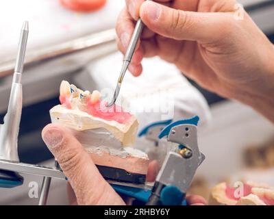 Dall'alto del raccolto, l'ortodontista maschile anonimo che fissa la dentatura sulla cera utilizzando una spatola dentale in acciaio inossidabile durante il lavoro in laboratorio Foto Stock