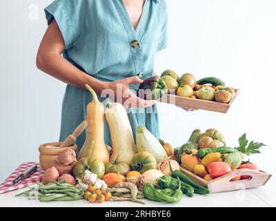 Coltiva una signora irriconoscibile in piedi vicino al tavolo con una pila di zucche e peperoni freschi maturi e verdure assortite, tenendo i pomodori contro il bianco Foto Stock