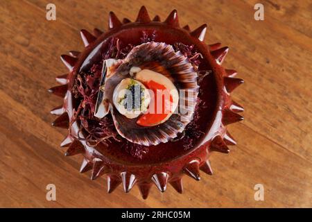 Vista dall'alto delle deliziose capesante fresche con caviale nero servite in una ciotola a forma di riccio di mare su un tavolo di legno in una stanza luminosa Foto Stock