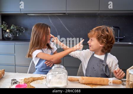 Fratello e sorella allegri che toccano il naso l'uno dell'altro con le dita indice sporche di farina mentre impastano l'impasto seduto al bancone della cucina Foto Stock