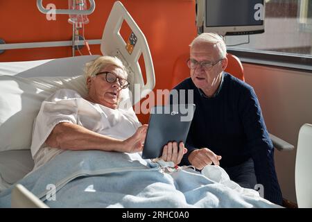 Paziente anziana con i capelli corti su occhiali sfoglia tablet e mostra il video al marito mentre è sdraiato sul divano medico durante la riabilitazione se Foto Stock
