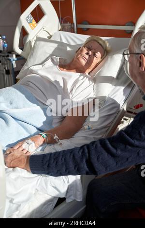 Dal lato superiore del raccolto uomo anziano che tiene la mano di moglie malata sdraiata sul lettino medico con catetere venoso collegato durante il processo di riabilitazione in mod Foto Stock