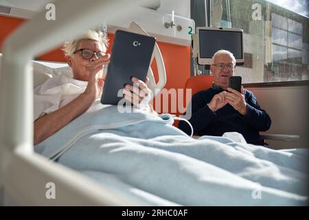 Uomo anziano serio seduto in poltrona e messaggi sullo smartphone vicino moglie malata sdraiato sul divano e utilizzando tablet durante la riabilitazione in ospedale Foto Stock