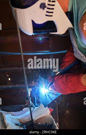 Vista laterale di un lavoratore maschile anonimo in maschera protettiva e guanti per la saldatura del metallo con torcia in fabbrica Foto Stock