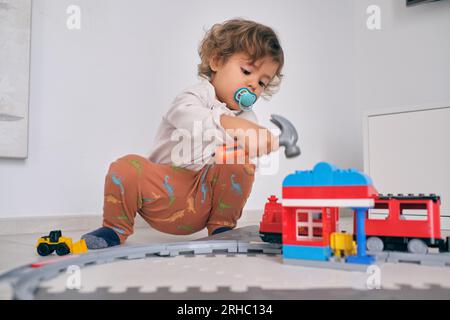 Tutto il corpo di un bambino curioso con un succhietto seduto sul pavimento in salotto e giocando con un martello giocattolo e la stazione ferroviaria a casa Foto Stock