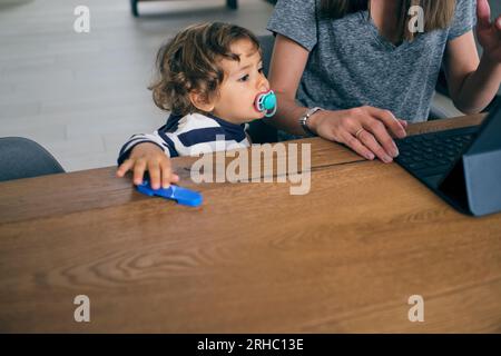 Angolo alto di bambino carino con succhietto per bambini seduto su un tavolo di legno e guardando lo schermo del tablet Foto Stock