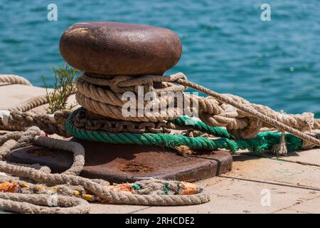Primo piano di un ormeggio in metallo arrugginito avvolto in spesse corde e catene sul marciapiede del porto in riva al mare. Foto Stock