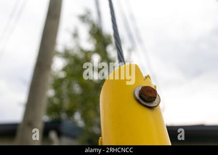 Primo piano di un palo metallico giallo con bullone e dado arrugginiti - cavi collegati - alberi e edificio sullo sfondo Foto Stock