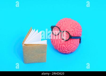 Simpatico personaggio del cervello umano adornato con occhiali, profondamente coinvolto nella lettura di un libro isolato su sfondo blu. Educazione e studio correlato concep Foto Stock