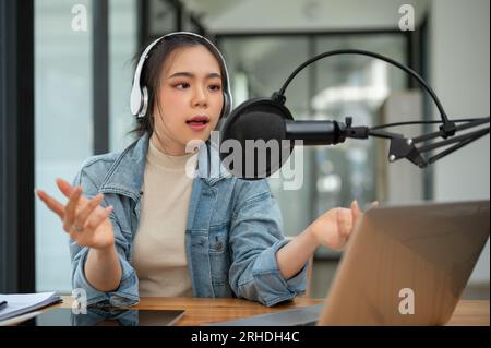 Un podcaster o conduttrice radiofonica asiatica professionale e focalizzata sta parlando in un microfono, annunciando notizie e trasmettendo il suo podcast audio dal vivo Foto Stock