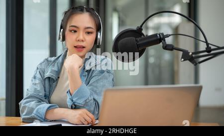 Una bella e professionale conduttrice radiofonica asiatica parla al microfono, annunciando o condividendo notizie interessanti con il suo pubblico online durante il corso Foto Stock