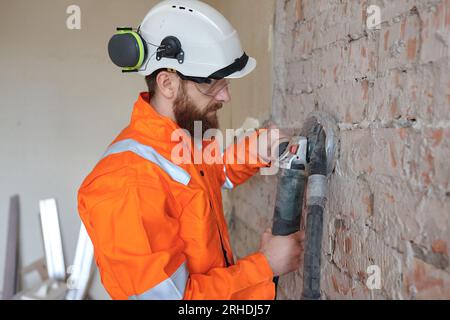 Operaio edile che esegue la rettifica di pareti in mattoni utilizzando una rettificatrice con aspirapolvere collegato. Foto Stock