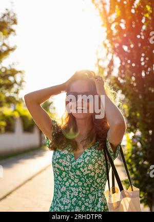 Giovane donna allegra con occhiali da sole e abito estivo in piedi per strada e sorridente davanti al tramonto con retroilluminazione. Foto Stock