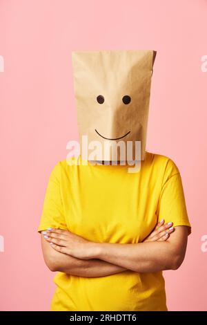 Giovane donna sicura di sé con un sorridente sacchetto di carta sulla testa e braccia incrociate su sfondo rosa. Concetto di emozioni Foto Stock