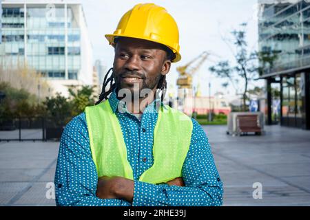 Capo ingegnere civile di etnia africana che indossa un casco di sicurezza in piedi all'aperto sorridente e guardante intorno sorride, poliziotto Foto Stock