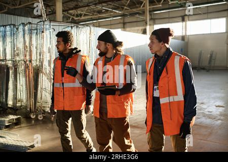 Tre giovani costruttori interculturali in giacche di sicurezza camminano lungo una spaziosa officina e discutono su quali nuove attrezzature dovrebbero essere acquistate Foto Stock