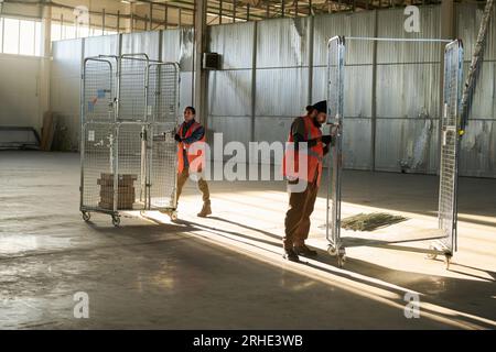 Foto lunga di due giovani ingegneri maschi impegnati nella spinta e riparazione di grandi carrelli metallici industriali mentre lavoravano in un magazzino spazioso Foto Stock