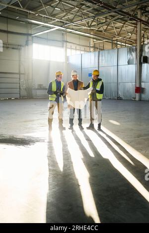 Foto lunga di tre giovani costruttori o ingegneri che discutono di progetto con il piano dell'edificio mentre si trovano in un magazzino illuminato dal sole Foto Stock