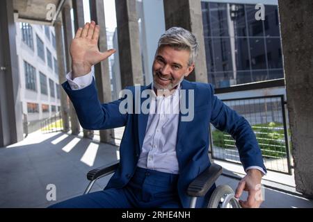 Uomo disabile dai capelli grigi che indossa una tuta ufficiale in sedia a rotelle, solleva il palmo, dà cinque in alto Foto Stock