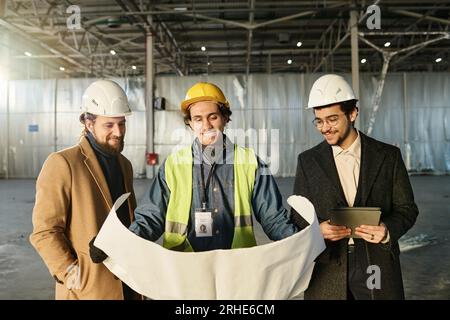 Gruppo di giovani architetti felici in caschi protettivi che esaminano lo schizzo della costruzione o il piano di costruzione su un progetto non arrotolato durante la riunione di lavoro Foto Stock