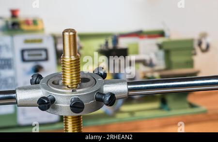 Filiera per la lavorazione dei metalli in acciaio in corrispondenza della filettatura della vite di taglio su un'asta in ottone. Utensile per filettatura manuale affilato nel giramaschi su sfondo sfocato dell'officina con tornio verde. Foto Stock