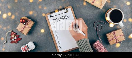 Buon anno 2024. La mano di una donna che scrive 2024 obiettivi nel notebook. Computer portatile, confezione regalo, tazza di caffè, decorazione natalizia. Vista dall'alto, base piatta. Banner i Foto Stock