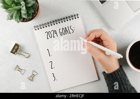 La mano di una donna che scrive 2024 obiettivi nel notebook. Computer portatile, impianto domestico, tazza di caffè su un tavolo leggero. Vista dall'alto, base piatta Foto Stock