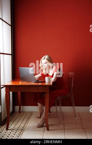 Giovane donna adulta seduta al tavolo e su cui lavora computer portatile Foto Stock