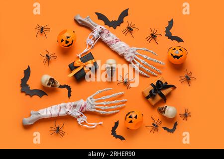 Composizione con mani scheletriche, confezioni regalo, teschi, pipistrelli di carta e ragni per festeggiare Halloween su sfondo arancione Foto Stock