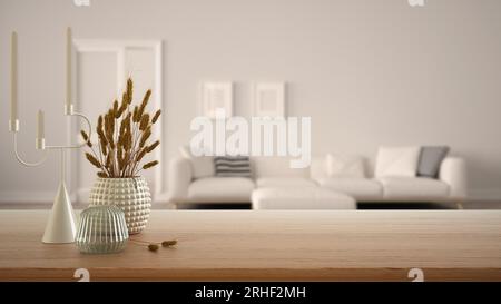 Tavolo in legno, scrivania o ripiano in primo piano con vasi in ceramica e vetro con piante secche, cannucce su vista sfocata del soggiorno bianco con divano e porta, m Foto Stock