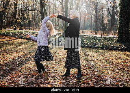 Madre spensierata che fa girare la sua figlia adolescente in autunno nel parco pubblico della città. Madre e figlia adolescente che giocano insieme nel parco autunnale Foto Stock