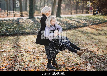 Madre spensierata che fa girare la sua figlia adolescente in autunno nel parco pubblico della città. Madre e figlia adolescente che giocano insieme nel parco autunnale Foto Stock