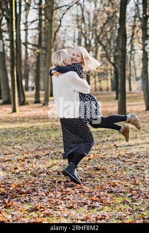 Madre spensierata che fa girare la sua figlia adolescente in autunno nel parco pubblico della città. Madre e figlia adolescente che giocano insieme nel parco autunnale Foto Stock