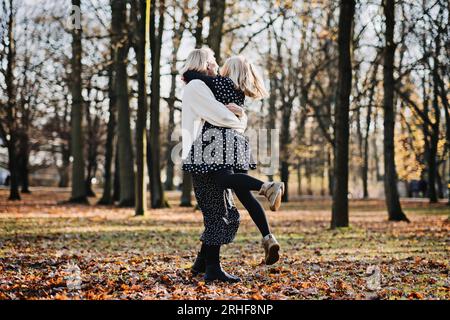 Madre spensierata che fa girare la sua figlia adolescente in autunno nel parco pubblico della città. Madre e figlia adolescente che giocano insieme nel parco autunnale Foto Stock