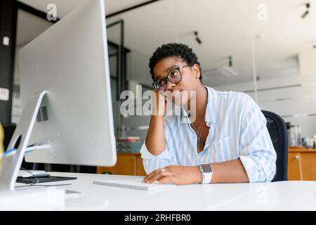 Studentessa africana stanca e annoiata o dipendente d'ufficio sentendo mancanza di energia e motivazione, svolgendo un lavoro monotono e noioso, seduto di fronte al pc, riflettendo sul cambiamento di lavoro, bruciando il concetto Foto Stock