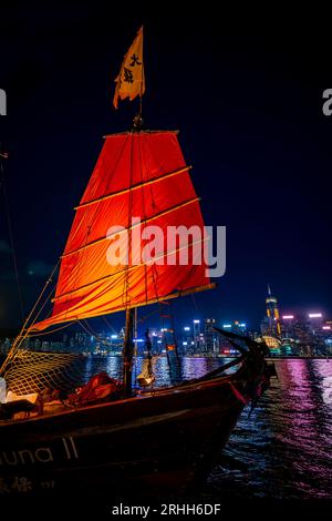 Fantastica vista notturna di una giunca luminosa nel Victoria Harbour di fronte al fantastico skyline di Hong Kong con i suoi impressionanti grattacieli Foto Stock