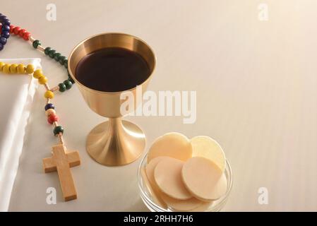 Coppa di calice con vino e ospiti consacrati in un contenitore su tavolo bianco e croce cristiana in legno. Vista elevata. Foto Stock