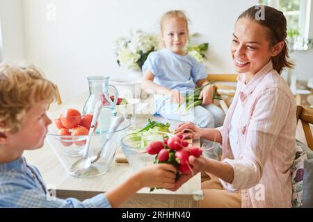 Vista laterale della madre felice che prende il ravanello dal figlio seduto sul tavolo a casa Foto Stock